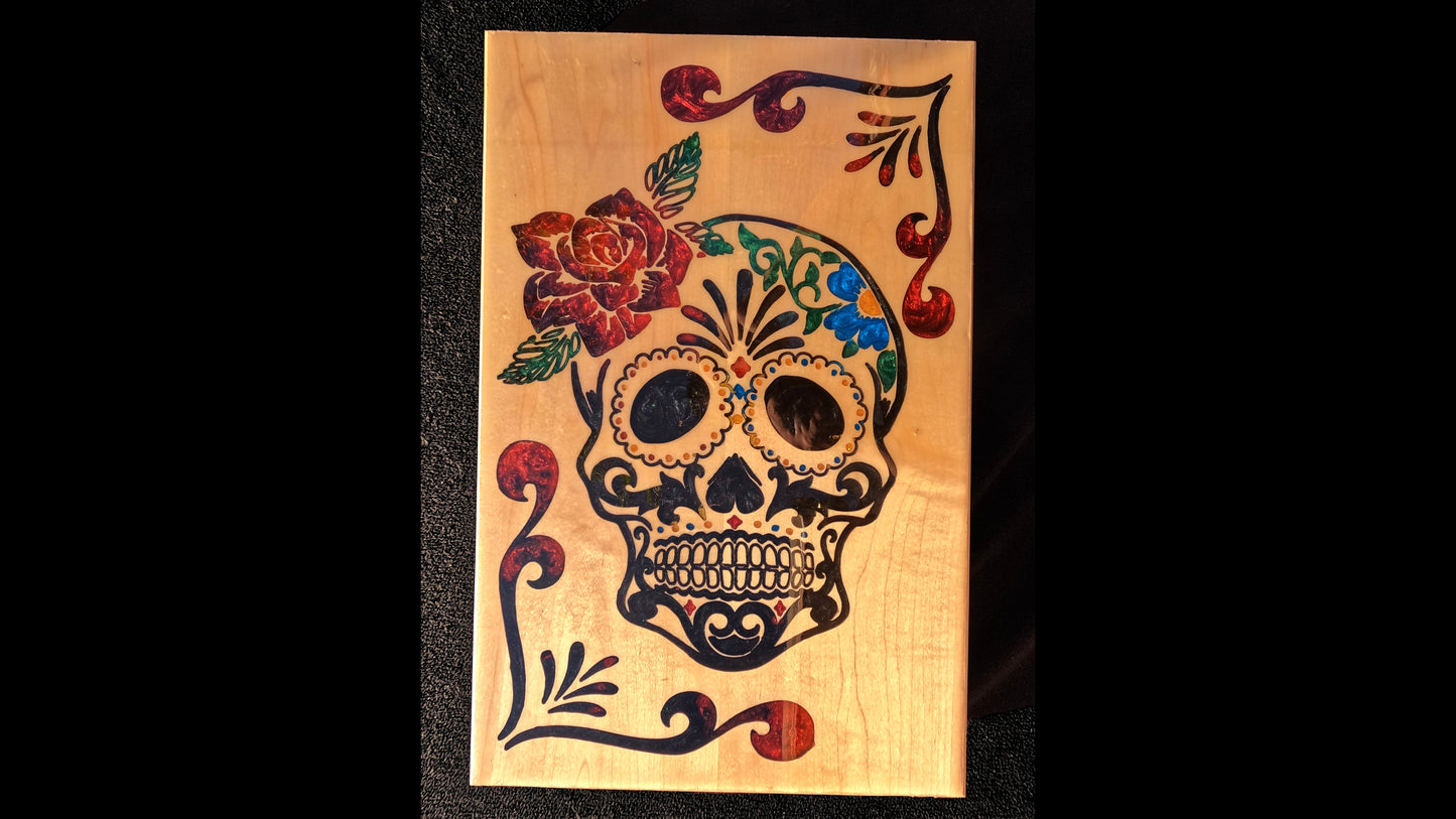 Dia de Los Muertos skull design as an epoxy inlay on a piece of hard maple wood. There are various colors of epoxy with a red rose on the skulls head, and black and red swirl designs in the corners.