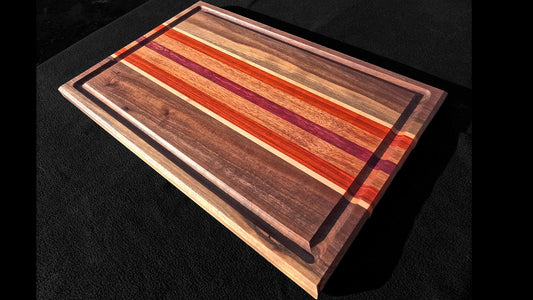 Front corner view of the Board to death stripped pattern Cutting board, featuring Walnut, maple, padauk, African mahogany, and purple heart hardwoods.